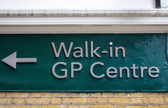 A turquoise sign with grey writing which reads walk-in GP centre with an arrow pointing left. It's on a yellow brick building